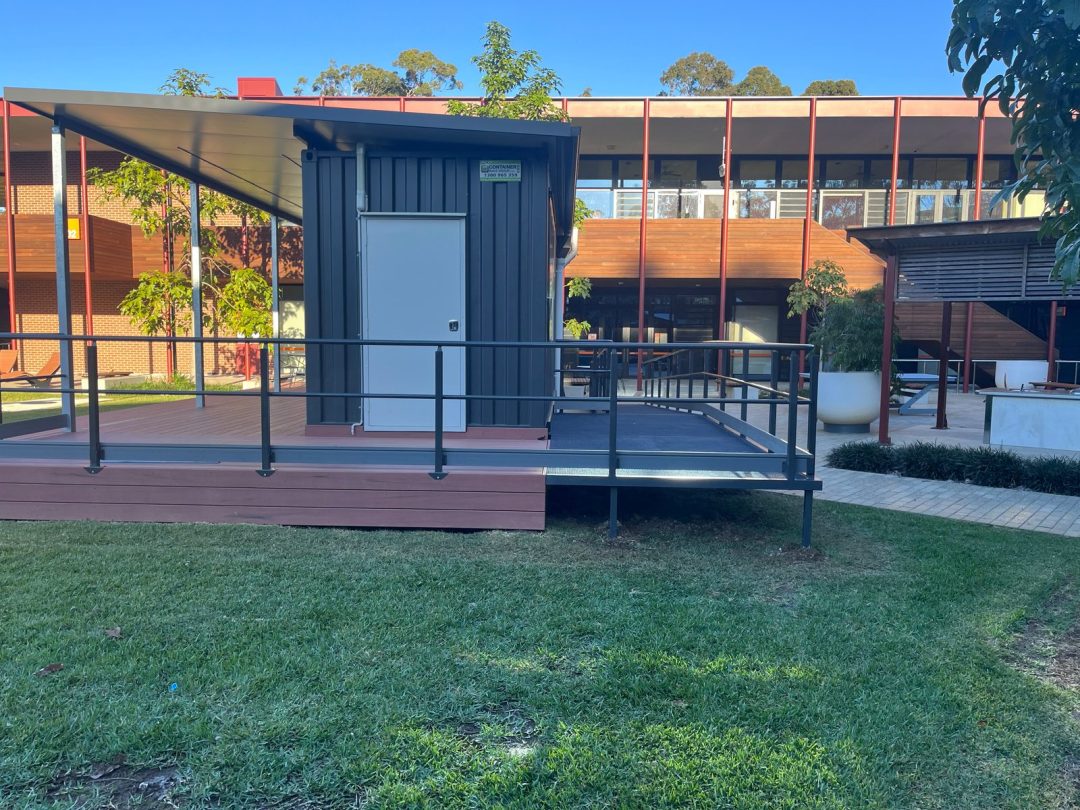 End of the dark container with a white door, the covered deck to one side and a railed ramp in front, on a lawn