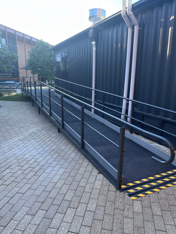 Long access ramp with handrails and a yellow and black warning strip at its base, running beside the dark container wall with downpipes and a rooftop flue