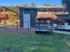 End of the dark container with a white door, the covered deck to one side and a railed ramp in front, on a lawn
