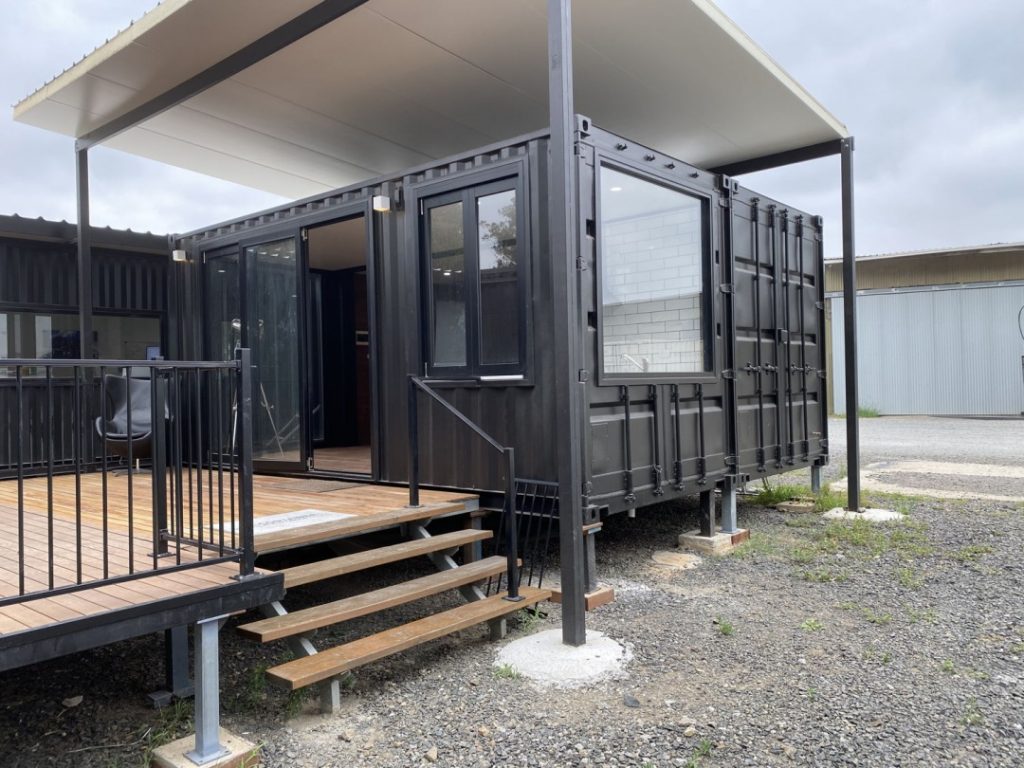 Container building on steel posts and concrete pad footings, with a raised timber deck and stairs