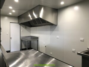Inside a 20ft commercial kitchen container: stainless steel range hood, benches and lined white walls